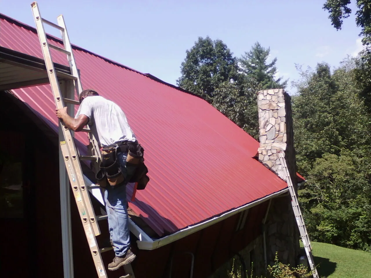 Roofer on a ladder beside a red metal roof and stone chimney — Soffit & Fascia Repair by Roofing Brothers in Simi Valley